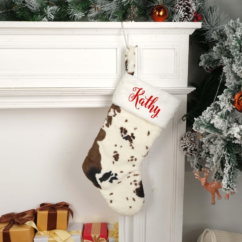 May include: A white and brown cow print Christmas stocking with a white faux fur cuff. The stocking is personalized with the name "Kathy" in red embroidery.