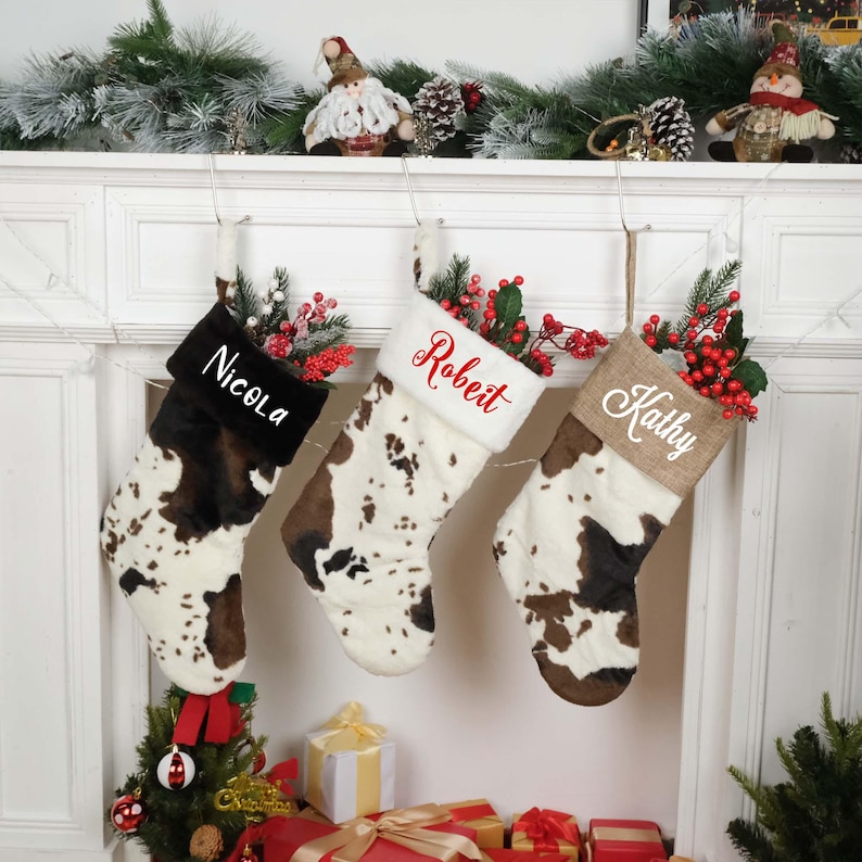 May include: Three personalized Christmas stockings with cowhide print fabric. The stockings are hung on a white fireplace mantel. The names Nicola, Robert, and Kathy are embroidered on the stockings in red.