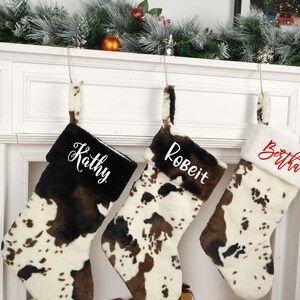 May include: Three personalized Christmas stockings with a cowhide pattern. The stockings are hung on a white fireplace mantel. The names Kathy, Robert, and Bertha are written in white on the stockings.