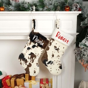May include: Two personalized Christmas stockings with a cow print design. The stocking on the left is labeled "Nicola" and the stocking on the right is labeled "Robert".