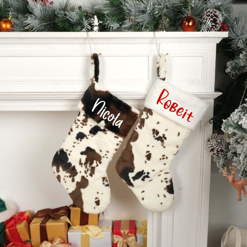 May include: Two personalized Christmas stockings with a cow print design. The stocking on the left is labeled "Nicola" and the stocking on the right is labeled "Robert".