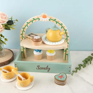 May include: A wooden toy tea set with a green and white stand, a yellow teapot, two yellow cups, two spoons, and various pastries. The stand has a sign that says "Sandy".