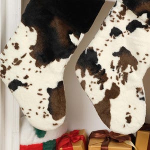 May include: Two brown and white cow print Christmas stockings hanging on a white shelf. The stockings are made of a soft, fuzzy material.