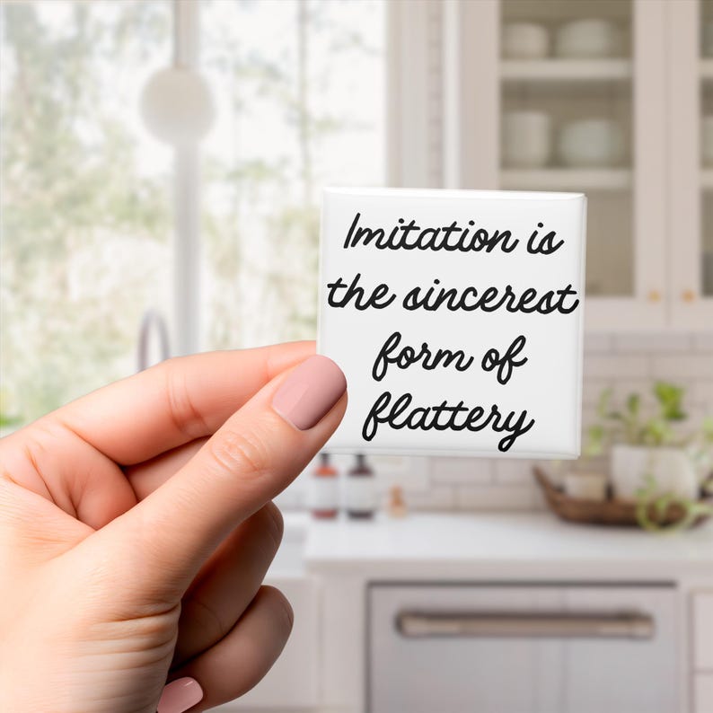 May include: A white square with the quote "Imitation is the sincerest form of flattery" in black script. The square is held in a hand with pink nail polish, against a blurred kitchen backdrop.