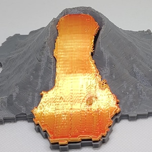 Puede incluir: Modelo impreso en 3D de un volcán en erupción. El volcán gris tiene una superficie texturizada, y la lava es de color naranja y amarillo vibrante. La base del volcán tiene una forma geométrica única.