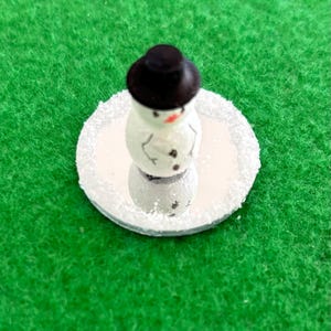 May include: A small snowman figurine with a black hat, a red nose, and black button eyes and buttons. The snowman is on a round, mirrored base with a white, glittery edge. The background is green.