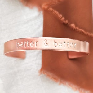 May include: A rose gold colored metal bracelet with the words "better & better" engraved on it.