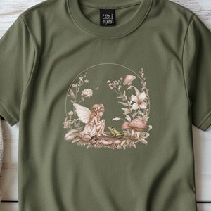 Könnte beinhalten: Ein grünes T-Shirt mit einem Aufdruck einer Fee, die in einem Wald mit einem Frosch sitzt. Das T-Shirt hat den Text "HOLI BYRD" auf dem Etikett.