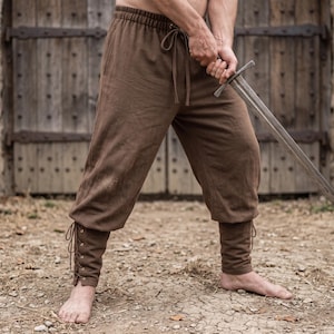 Può includere: Pantaloni marroni con elastico in vita e coulisse, tasche laterali e sezioni inferiori delle gambe allacciate con occhielli in metallo. I pantaloni sono indossati con una spada in metallo tenuta in mano, mostrando una vestibilità ampia.