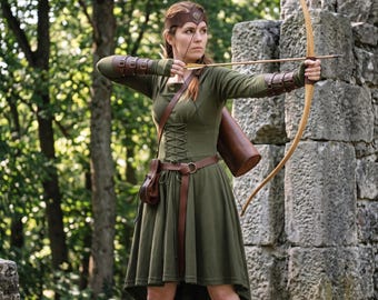 May include: An olive green dress with a lace-up bodice, worn with brown leather arm bracers, a matching headband, a belt with a small pouch, and a quiver. The outfit is paired with a wooden recurve bow and an arrow, creating a complete archer ensemble.