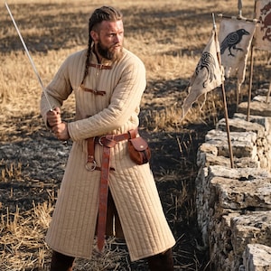 May include: A beige quilted long-sleeved tunic featuring vertical stitching, a high collar, and brown leather straps with buckles. A brown leather belt with an embossed pattern, a metal ring, and a small attached leather pouch are worn at the waist.
