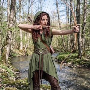 May include: A hooded olive green tunic with a high-low hemline, worn with a brown leather belt featuring two silver rings and long dangling straps. The outfit includes brown trousers, a wooden recurve bow, and a quiver filled with arrows.