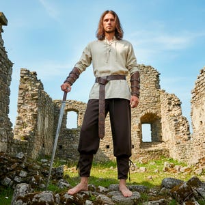 Op de afbeelding: Een middeleeuws geïnspireerde outfit met een beige shirt met lange mouwen, een bruine broek en een bruine riem met patroon. De outfit bevat leren armbeschermers en een zwaard. De persoon staat voor een stenen ruïne.