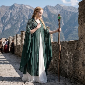 May include: A floor-length white dress layered with a sheer, dark green hooded cape featuring delicate silver bead embellishments. The garment is paired with a tall, twisted wooden staff topped with a polished, vibrant green gemstone.