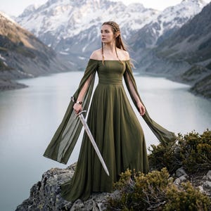 Può includere: Un abito lungo color verde oliva con spalle scoperte, maniche lunghe e fluide con spacchi e vita aderente. Il capo è indossato con una spada metallica tenuta in mano, caratterizzata da una lama appuntita e un'elsa a forma di croce.