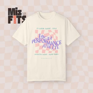Pode incluir: Uma camiseta branca com um design gráfico rosa e roxo que diz "It's real sweat. I'm a High Performance Athlete. Athletes sweat. Sweat baby. Miniha ay ay."
