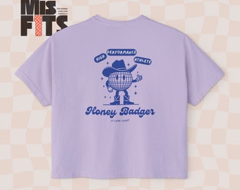 Puede incluir: Camiseta morada con un gráfico azul de un personaje de dibujos animados con un sombrero de vaquero y sosteniendo una bola de discoteca. El texto "Honey Badger" está escrito en una fuente retro debajo del personaje. El texto "It's Real Sweat!" está escrito en una fuente más pequeña debajo del texto principal.