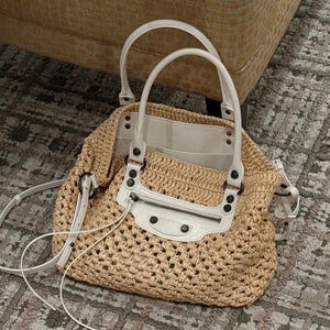 May include: A beige woven handbag with white leather accents. The bag features a top zipper, two handles, and a detachable shoulder strap. The bag is made of a woven material and has a white leather trim.
