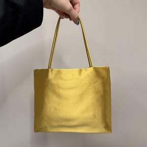 May include: A rectangular handbag in a solid mustard yellow color with a smooth, satin-like finish. The bag features two thin, matching yellow shoulder straps attached to the top edge, creating a simple, structured silhouette for carrying items.