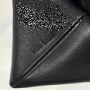 以下が含まれることがあります： 黒いレザー素材のアイテムで、表面には質感があり、斜めの縫い目と「YUU GOM」というエンボス加工の文字が施されています。素材は細かい粒状のパターンを示しており、2つのレザーセクションが鋭角に交わる部分にははっきりとした折り目が見られます。