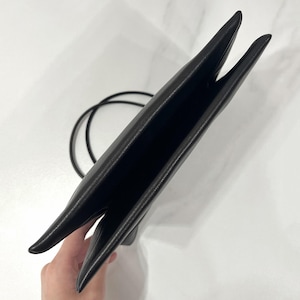 以下が含まれることがあります： 滑らかでマットな質感の黒いフェイクレザー製ショルダーバッグ。スリムな長方形のシルエットで、上部が大きく開いたデザインです。両サイドから伸びる細い黒いコードストラップが付いており、肩にかけて持ち運ぶことができます。