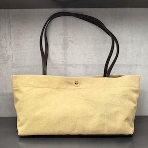 Puede incluir: Un bolso tote rectangular hecho de tela de lona de color beige claro con dos correas de hombro de cuero marrón oscuro. El bolso cuenta con un único botón de presión de metal dorado centrado en el borde superior de la abertura principal.