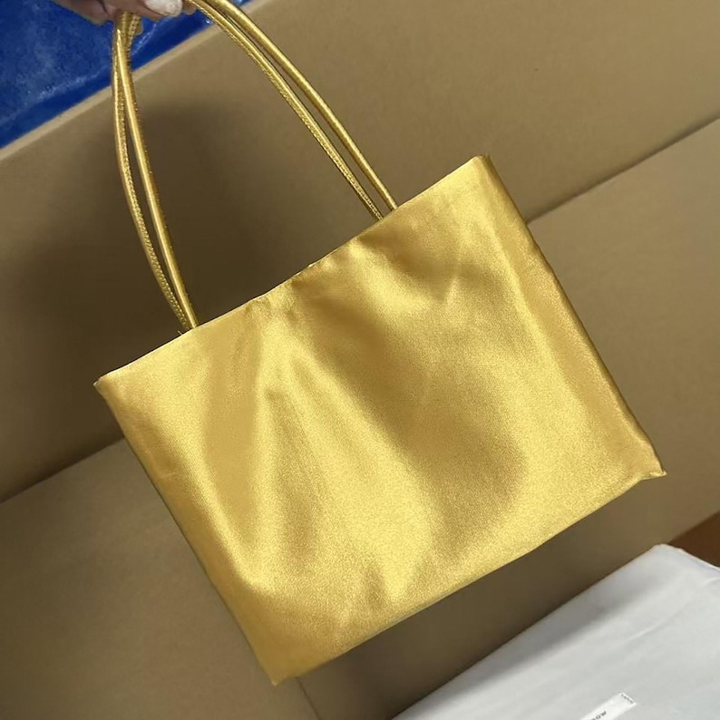May include: A rectangular tote bag in a metallic gold finish with a smooth, satin-like texture. The bag features two thin, matching gold shoulder straps attached to the top edge, creating a simple, structured silhouette with a soft sheen.