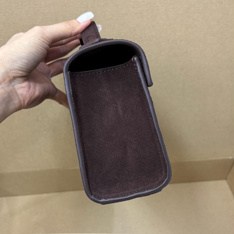 May include: A rectangular, dark brown suede handbag with a flap closure. The bag has a handle and a contrasting brown trim. The interior is visible, revealing a dark space. The bag is held by a hand.