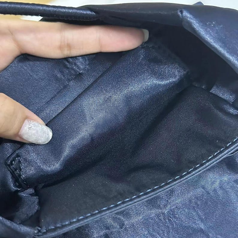May include: A dark navy blue satin handbag interior featuring a smooth, lustrous fabric lining. The bag has a structured opening with visible stitching along the edges and a deep, open compartment designed for storage, showcasing the fabric texture.