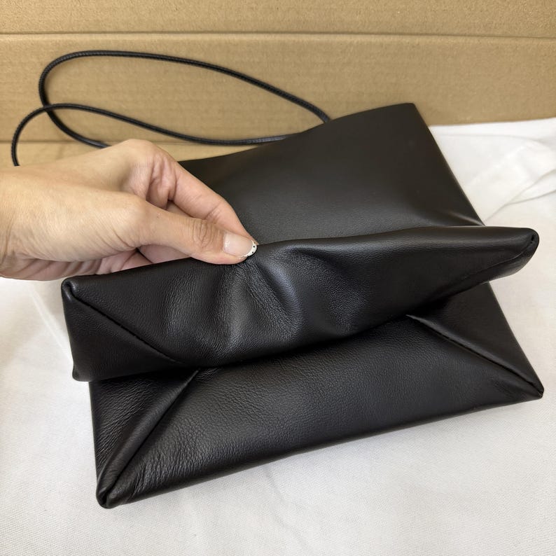以下が含まれることがあります： 柔らかくしなやかな質感の黒いレザーショルダーバッグで、細い同色のストラップが付いています。バッグは長方形で、折り畳まれたギャザー入りのエッジが特徴的で、前面に独特の層状の外観を作り出しています。