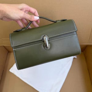 May include: Olive green handbag with a top handle and a silver-tone clasp. The rectangular bag features white stitching and a flap closure. A white dust bag is visible inside the open box.