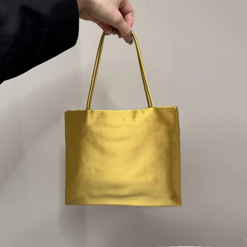 May include: A rectangular handbag in a solid mustard yellow color with a smooth, satin-like finish. The bag features two thin, matching yellow shoulder straps attached to the top edge, creating a simple, structured silhouette for carrying items.
