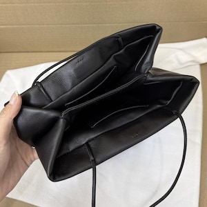 以下が含まれることがあります： 柔らかく構造化されていないシルエットの黒いレザーハンドバッグ。上部は広く開いており、内部には中央の仕切りによって作られた2つのメインコンパートメントがあります。バッグの両サイドには細い丸型のショルダーストラップが付いています。