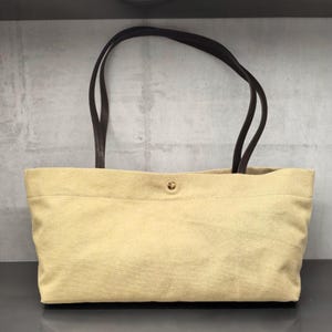 May include: A rectangular tote bag made of light beige canvas fabric. The bag features two dark brown shoulder straps attached to the top edge and a single gold-toned metal snap button closure centred at the top opening of the bag.