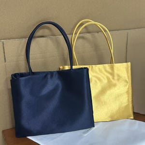 May include: Two rectangular handbags with thin shoulder straps. One bag is a deep navy blue, and the other is a bright metallic gold. Both bags feature a smooth, satin-like finish and a simple, structured design with a flat base and top opening.