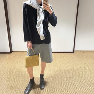 May include: A black long-sleeved shirt with a light gray sweater draped over the shoulders, paired with gray shorts, black socks, and dark shoes. A small, rectangular mustard yellow handbag with thin straps is held in one hand.