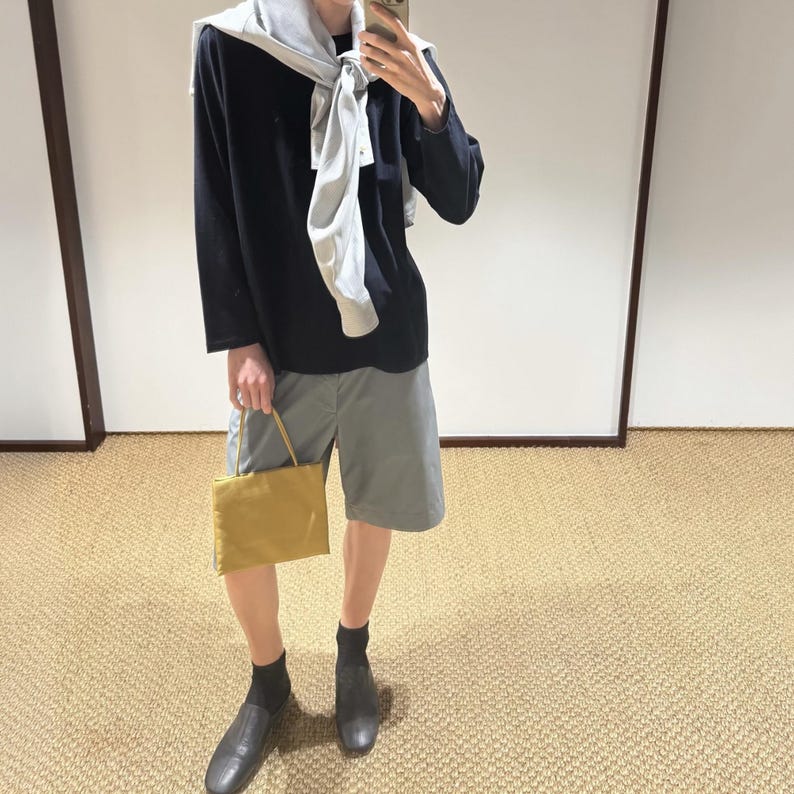May include: A black long-sleeved shirt with a light gray sweater draped over the shoulders, paired with gray shorts, black socks, and dark shoes. A small, rectangular mustard yellow handbag with thin straps is held in one hand.
