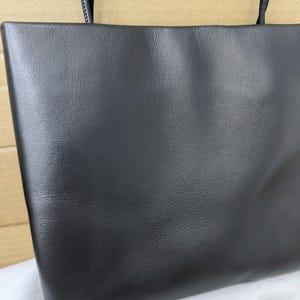 以下が含まれることがあります： 滑らかでわずかに質感のある表面を持つ黒いレザーのトートバッグで、細い黒のショルダーストラップが付いています。バッグは長方形で、端はまっすぐで清潔感があり、前面全体に均一なマット仕上げが施されています。