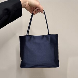 May include: A dark navy blue handbag with a smooth, satin-like finish. The bag features a rectangular body and two thin, matching navy blue top handles. The design is simple and structured, with a clean, solid-colored surface throughout.