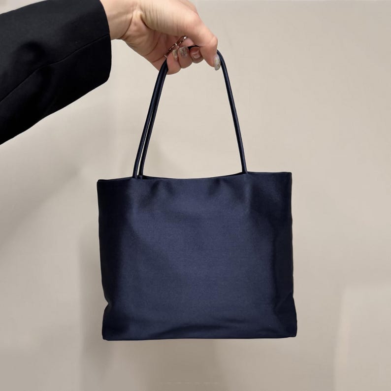May include: A dark navy blue handbag with a smooth, satin-like finish. The bag features a rectangular body and two thin, matching navy blue top handles. The design is simple and structured, with a clean, solid-colored surface throughout.