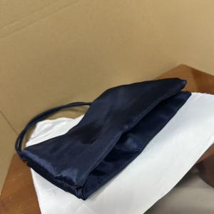 May include: A dark navy blue satin handbag with a smooth, lustrous finish. The bag features a rectangular, folded silhouette with a thin, matching navy blue shoulder strap attached to the side, presenting a clean and simple design structure.