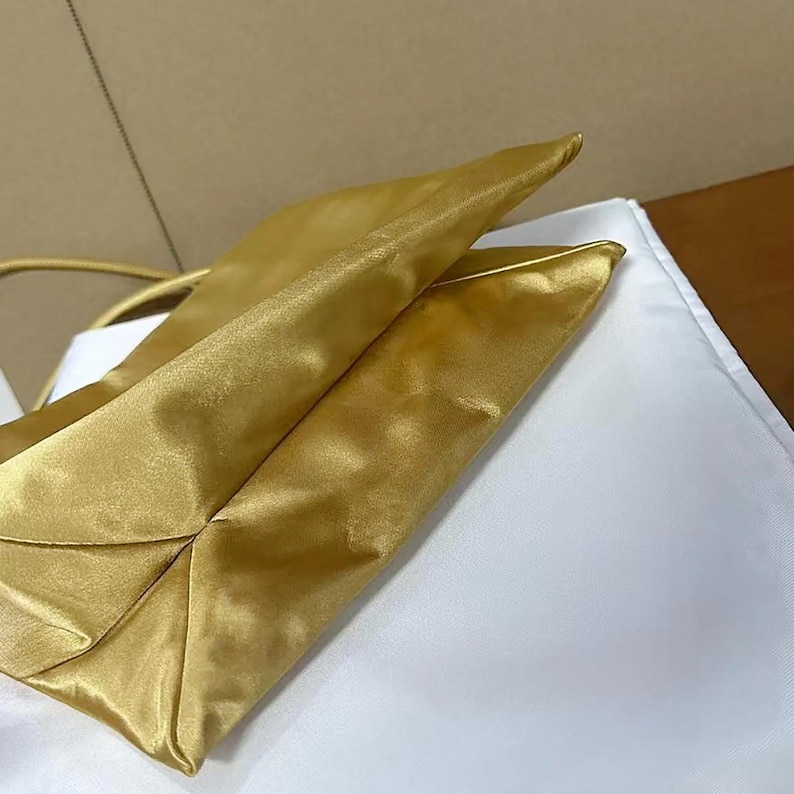 May include: A metallic gold handbag with a smooth, lustrous finish and a structured, folded design. The bag features a thin, matching gold strap and a clean, minimalist silhouette with visible seams along the edges, showcasing a reflective surface.