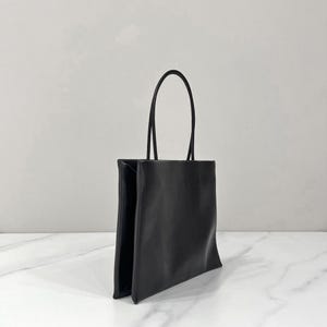 以下が含まれることがあります： 長方形のシルエットと滑らかでマットな質感を持つ黒いトートバッグ。バッグの上端には2本の細い垂直なハンドルが付いており、シンプルで機能的なこのアクセサリーに、すっきりと整った外観を与えています。