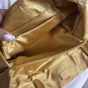May include: A gold satin handbag interior featuring a smooth, lustrous fabric lining with visible stitched seams and a rectangular pocket compartment. The bag has a soft, flexible structure with a consistent golden hue throughout the inner lining.