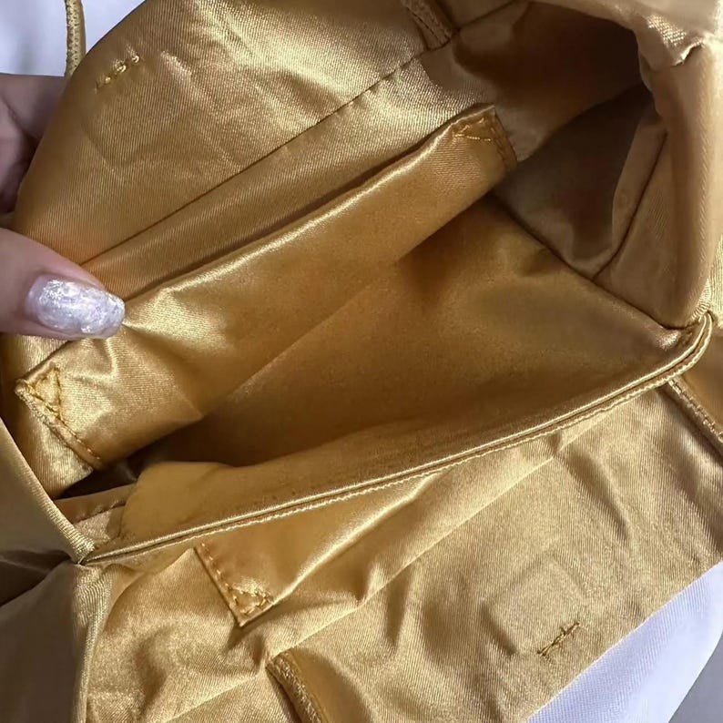 May include: A gold satin handbag interior featuring a smooth, lustrous fabric lining with visible stitched seams and a rectangular pocket compartment. The bag has a soft, flexible structure with a consistent golden hue throughout the inner lining.