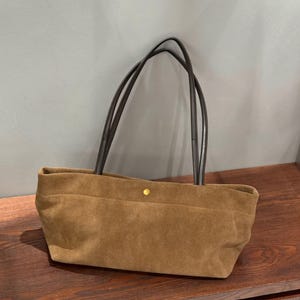 May include: A tan suede tote bag featuring a rectangular silhouette with a soft, textured finish. The bag includes two long, dark brown shoulder straps and a single small, centered gold-tone metal snap button closure at the top opening.