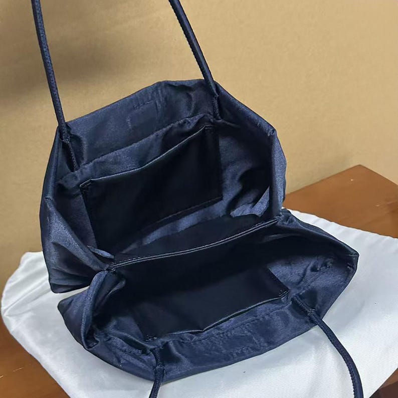 May include: A dark navy blue satin handbag with a structured open top, thin shoulder straps, and a smooth fabric interior. The bag features a rectangular internal pocket and a deep main compartment, showcasing a clean, minimalist design.