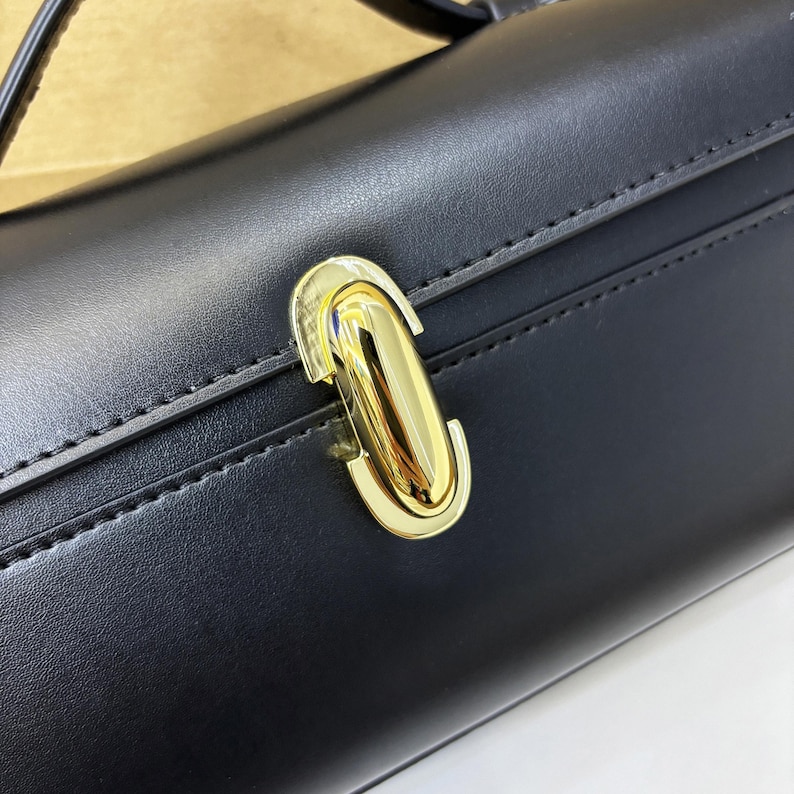 May include: Close-up of a black handbag with a gold-colored clasp. The bag has a rectangular shape and a smooth, leather-like texture. The clasp is a shiny, elongated oval shape.