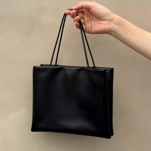 以下が含まれることがあります： 滑らかでマットな質感の無地の黒い長方形のハンドバッグ。バッグの上端には2本の細い垂直な黒いストラップが付いており、身の回りの物を持ち運ぶためのシンプルで構造的なデザインになっています。