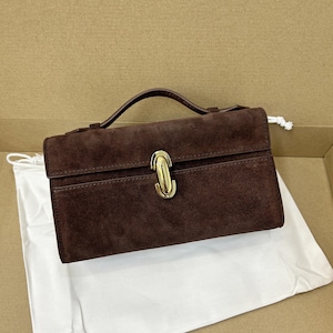 May include: A brown suede handbag with a top handle and a gold-colored clasp. The bag has a rectangular shape and a flap closure. A white drawstring bag is visible underneath the handbag.